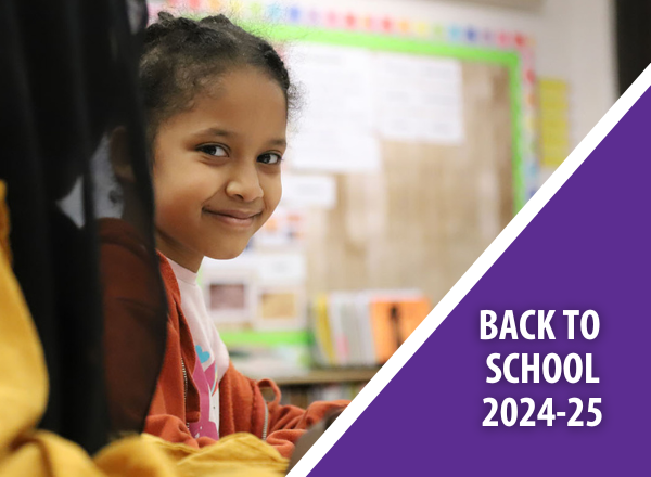 Smiling African-American student in an Elementary classroom writing at her desk w/ "Back to School 2024-25" in the corner.